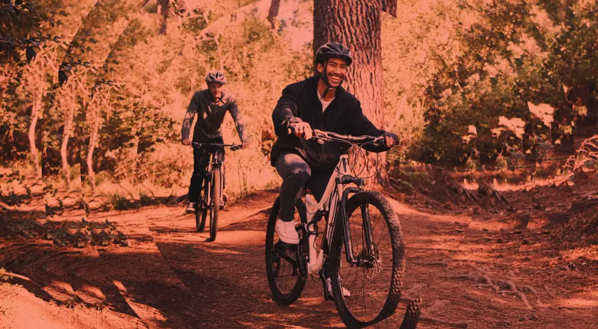 Deux cyclistes descendent une piste forestière en terre, entourés d'arbres. L'avant-plan montre un cycliste souriant, portant un casque et des vêtements de sport. Un autre cycliste suit derrière.