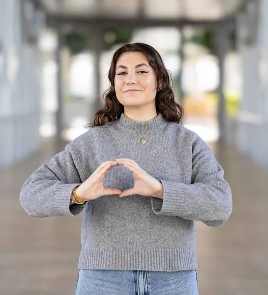 Usměvavá žena v šedém svetru a džínách před moderní budovou, ruce tvoří logo OneHealth.
