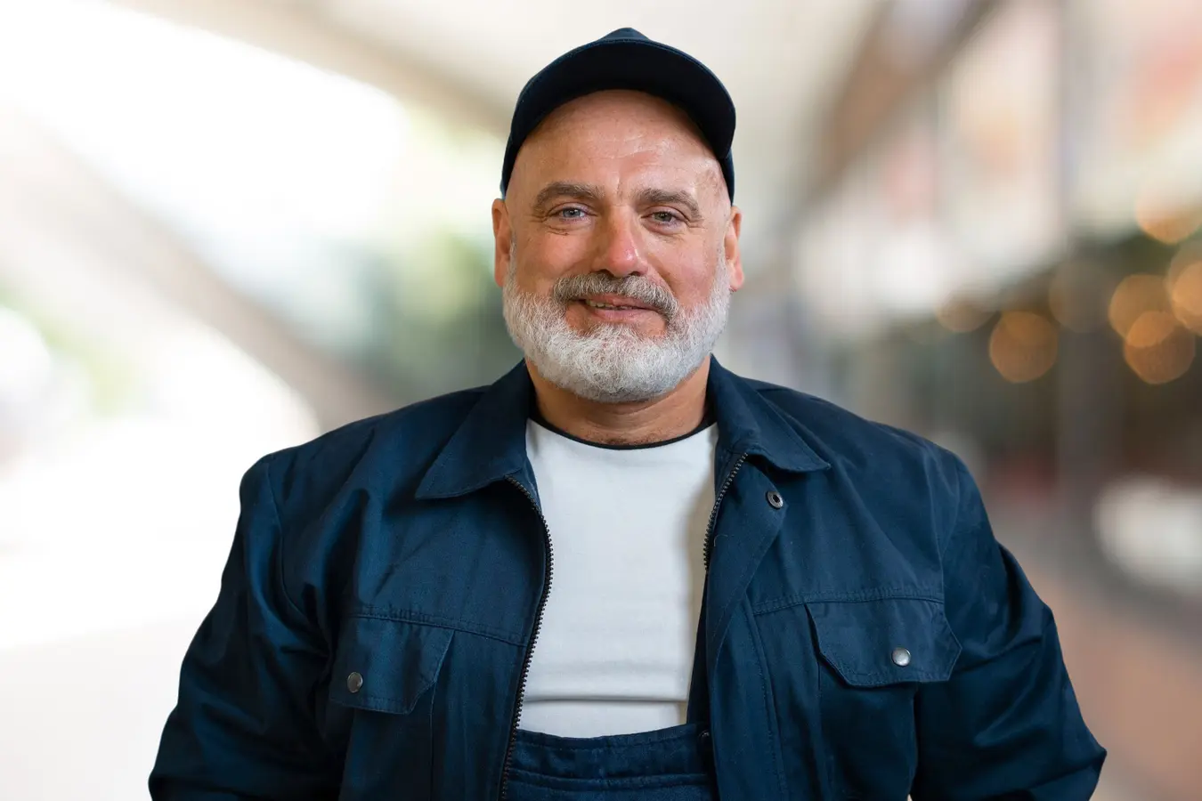 Lächelnder Mann mit grauem Bart, schwarzer Kappe, blauer Jacke und weißem Hemd.