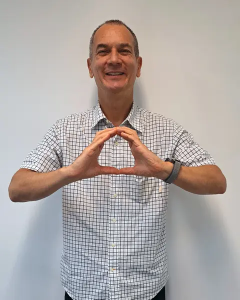 Homem sorridente formando o logotipo da OneHealth com as mãos, vestindo uma camisa xadrez.